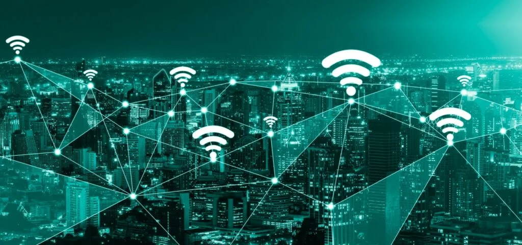 Futuristic image depicting a network of connections over a cityscape at night, highlighted in vibrant teal tones. The scene includes multiple WiFi symbols rising above the skyline, illustrating the concept of widespread digital connectivity and communication. This image metaphorically represents the pervasive and interconnected nature of mobile and desktop traffic on the internet, relevant to discussions on optimizing website traffic for various devices.