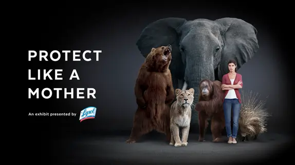 Advertisement with a bear, lion, elephant, and a woman standing side by side under the spotlight with the text ‘PROTECT LIKE A MOTHER’ above. The Lysol logo and ‘An exhibit presented by’ text are visible in the bottom left corner, symbolizing guardianship and safety.