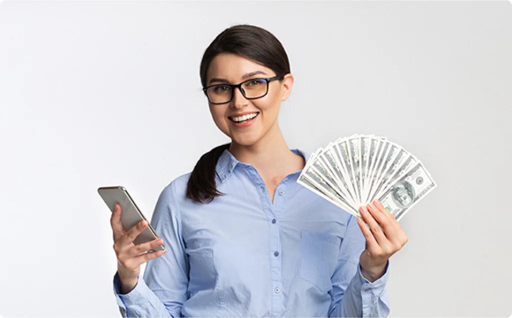 A professional woman with dark hair, wearing glasses and a blue button-up shirt, confidently holds a smartphone in one hand and a fan of cash in the other. Her bright smile suggests success and satisfaction, symbolizing effective monetization strategies through website advertising that have led to increased income