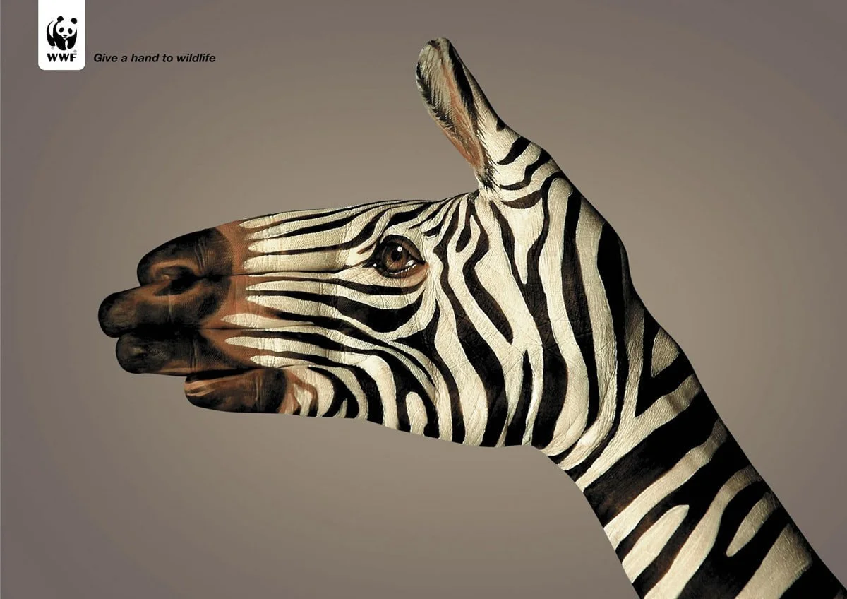 This creative advertisement from WWF features a human hand painted to resemble a zebra’s head and neck, raised as though it is the zebra itself. The hand's position and the painting align to form a seamless image of the zebra's profile, against a muted brown background. At the top left, the WWF logo with a panda is visible, and beside it, the text 'Give a hand to wildlife' encourages conservation efforts. The visual metaphor suggests that human action is crucial in protecting wildlife, as symbolized by the hand forming the essence of the zebra.
