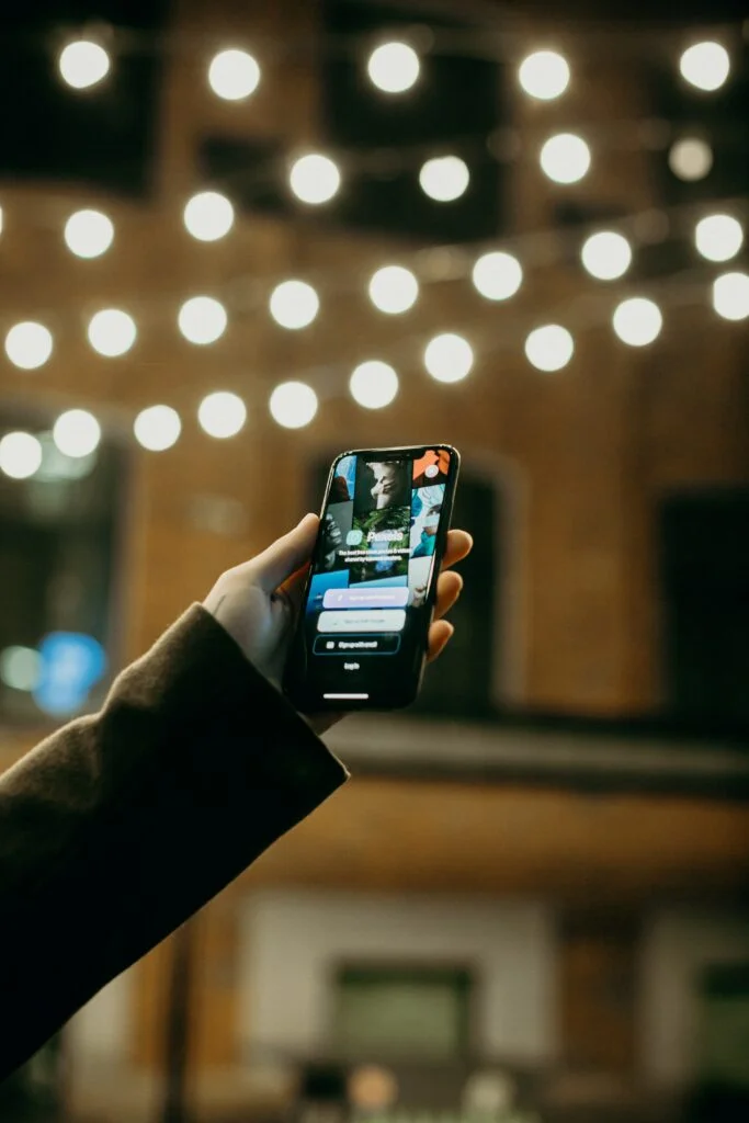 The image displays a person's hand holding a smartphone with a visible screen. The phone is illuminated by a bokeh background of large, out-of-focus light bulbs, suggesting an indoor setting, possibly a café or an event space. The screen shows a social media or content streaming application with images and user interface elements, such as icons and text, indicative of an active engagement or browsing. The overall scene captures a moment of digital interaction in a modern, connected environment, highlighting the ubiquity of mobile devices in accessing online content.