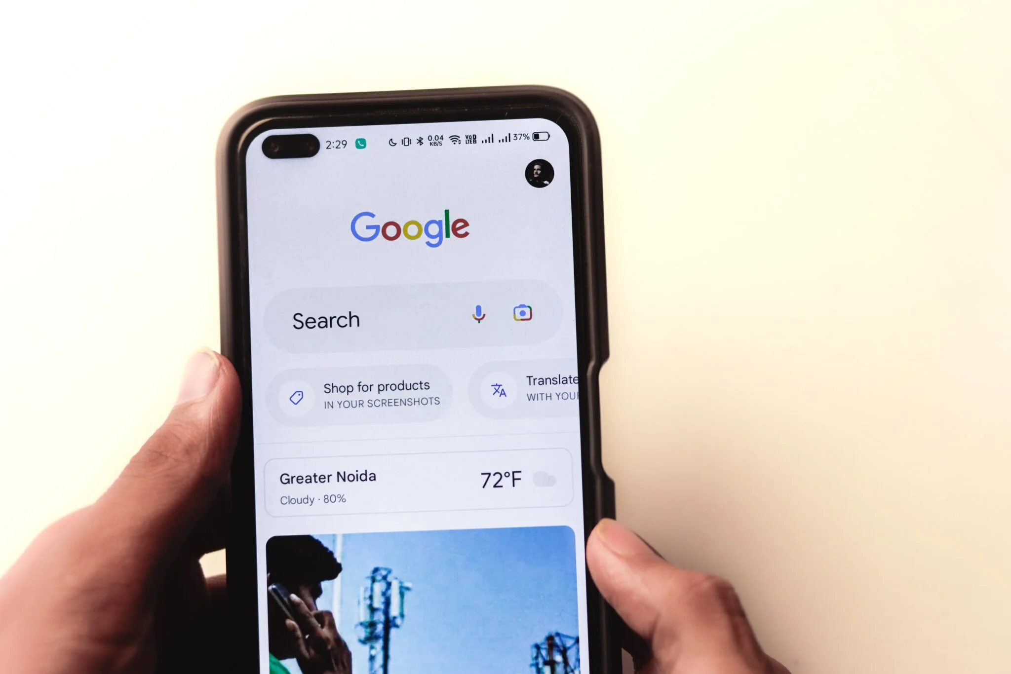 A person holding a smartphone displaying the Google search homepage, with options like ‘Search’, ‘Shop for products’, and ‘Translate’ visible on the screen, along with weather information for Greater Noida.