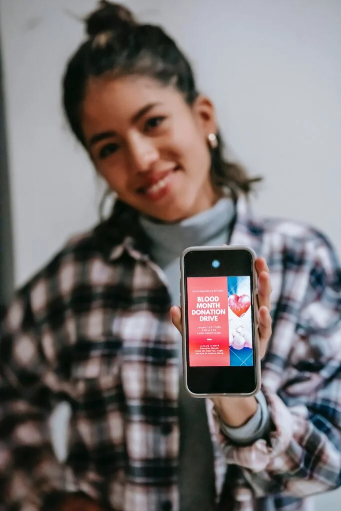 A person in a flannel shirt is holding a smartphone that displays an advertisement for a ‘Blood Month Donation Drive’, with the face obscured for privacy. The ad features an image of red and white flowers with the campaign title prominently displayed