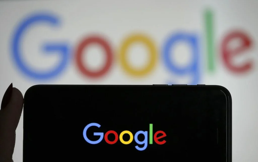 A silhouette of a person’s hand holding up a smartphone against a blurred background displaying the colorful Google logo. The phone's screen also shows a clear, focused Google logo, creating a visual echo effect and highlighting the brand's presence in both the digital and physical space