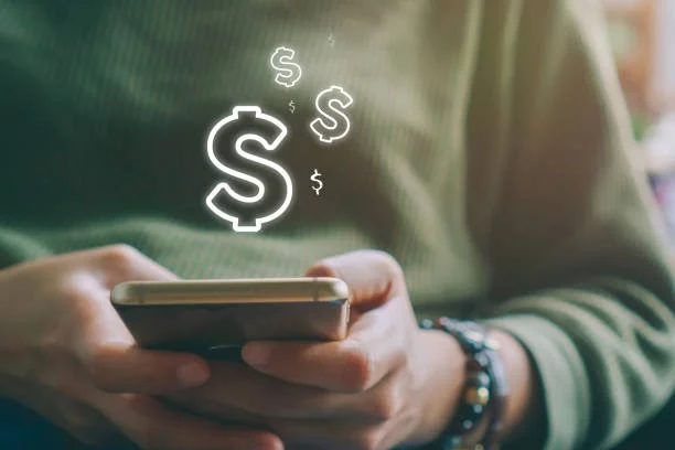 A person wearing a green top holds a smartphone horizontally. The hands are visible, one supporting the bottom of the phone while the other steadies it from the side. Above the smartphone, three glowing white dollar signs of varying sizes suggest increasing earnings. The blurred indoor background adds a sense of natural light. Overall, the image symbolizes the potential for online earnings through AdSense.