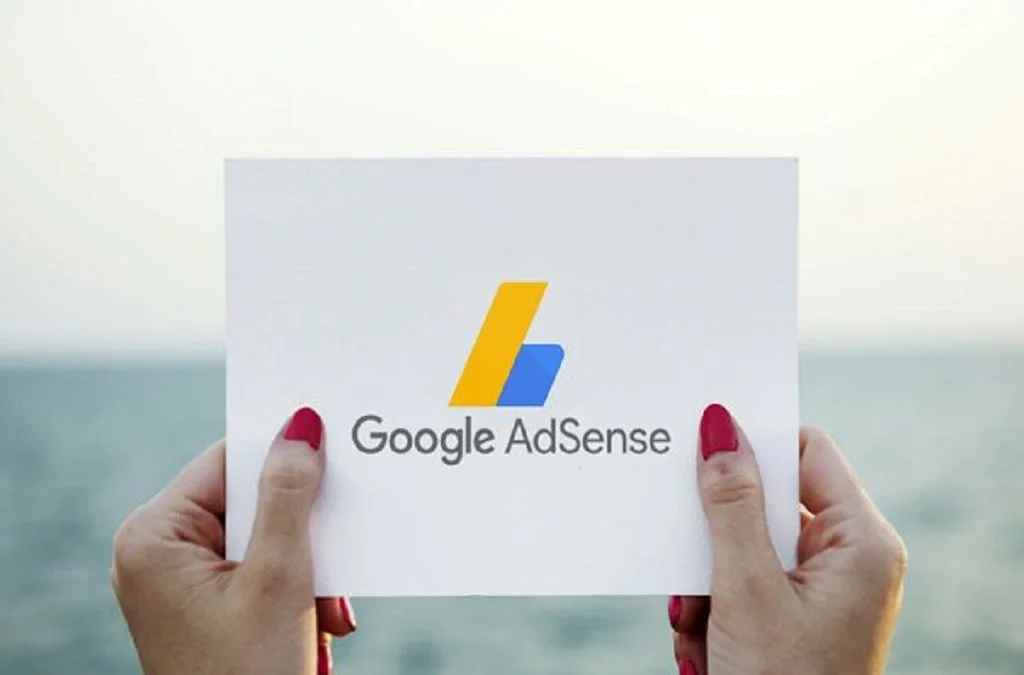 A person holding a white card with the Google AdSense logo and text, against a serene seascape background with soft lighting, suggesting a peaceful setting.