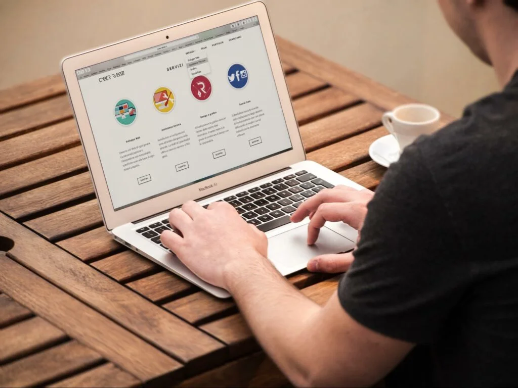 An individual is seen interacting with a laptop on a wooden table, which displays a webpage with indistinct colorful icons and text. A white coffee cup rests beside the laptop, alluding to a casual work environment aimed at enhancing website engagement