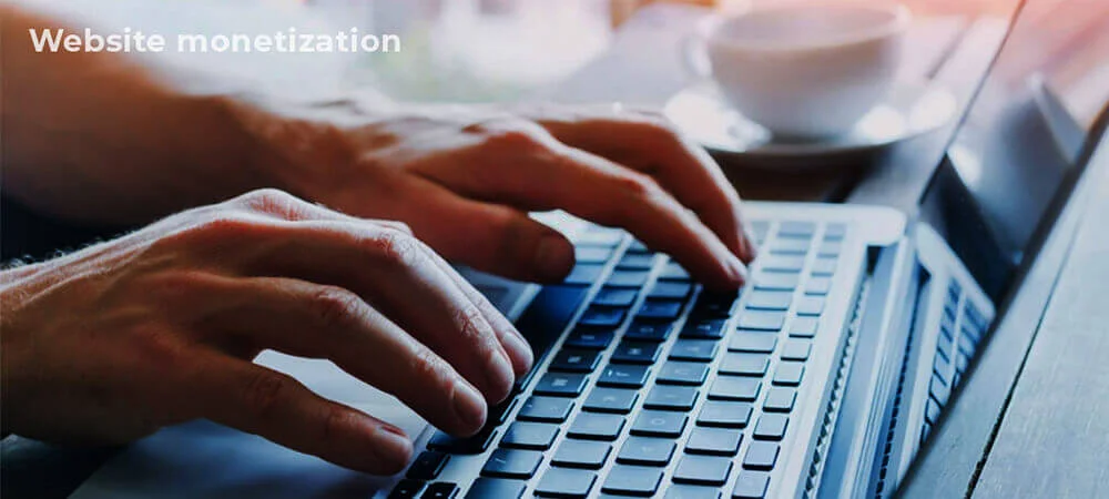 The image depicts a pair of hands typing on a laptop keyboard, with the text “Website monetization” displayed at the top left corner. In the background, a white coffee cup sits on a saucer, suggesting a work or productivity setting. The overall mood conveys focus and productivity
