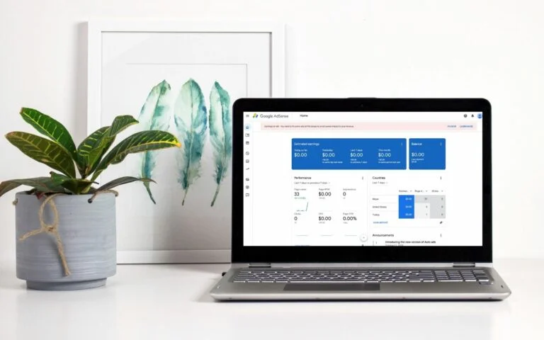 A laptop displaying Google AdSense dashboard with analytics data is placed on a white surface. To the left, there’s a potted plant with green leaves and a framed artwork of abstract green brush strokes hanging on the wall in the background