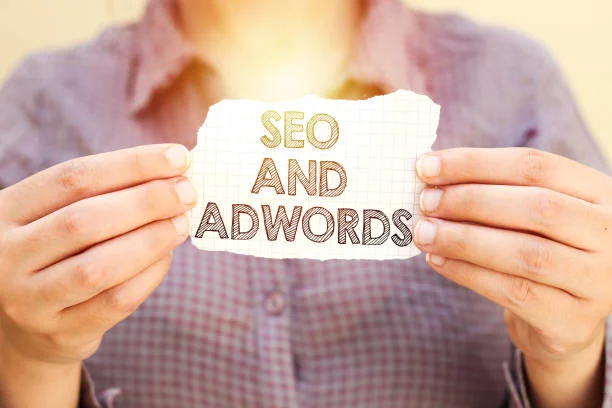 A close-up photo of two hands holding a piece of white paper with the words 'SEO AND ADWORDS' handwritten in what appears to be pencil. The paper is torn around the edges, giving it a rough appearance. The background is a warm, blurred glow, possibly suggesting the idea of enlightenment or discovery in the context of search engine optimization and online advertising strategies as part of effective website monetization following AdSense policies.