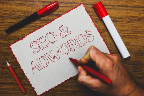 A person's hand is writing the words 'SEO & ADWORDS' in red on a piece of rustic, torn-edged paper. Above the paper, there's a closed red pen and a pencil, and to the right, a red marker with its cap off. The hand holds a red colored pencil, indicating the process of working on search engine optimization and Google AdWords strategies. This image represents the meticulous planning and attention to detail required for the successful and policy-compliant monetization of a website