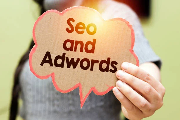 A person holds a speech bubble-shaped cardboard sign that reads 'SEO and Adwords' in bold letters, symbolizing the discussion and implementation of search engine optimization and online advertising strategies. The person is out of focus in the background, suggesting that the message is the focal point. This visual metaphorically emphasizes the importance of these digital tools for maximizing online earnings through platforms like AdSense Media.