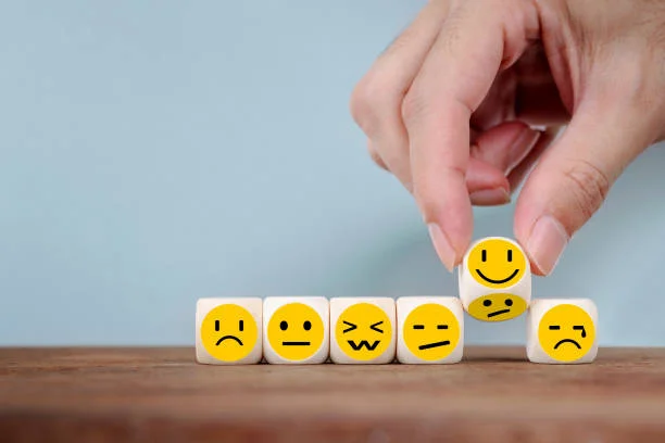 A hand is adjusting small cubes with various emoji faces on them, changing from a frowning expression to a smiling one. This represents the concept of influencing customer satisfaction or mood. The sequence of emojis shows a progression from negative to positive emotions, symbolizing the improvement of customer experience. The background is a simple, soft blue, providing a calm and neutral setting