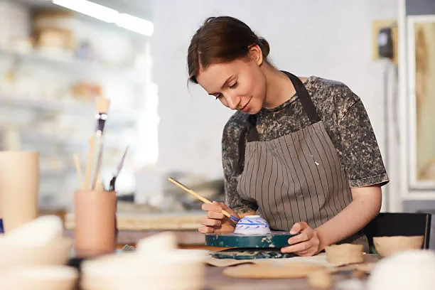 A person in a patterned shirt and apron is painting pottery in a well-lit workshop, surrounded by various art supplies