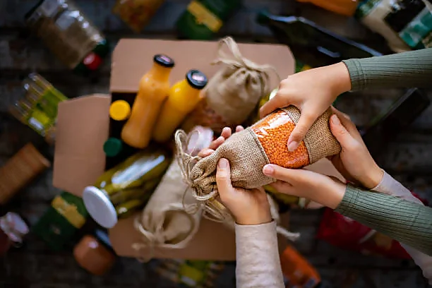 An adult and a child’s hands holding a burlap sack filled with orange lentils, surrounded by various food items including bottles of juice, jars, and packaged goods on a patterned floor