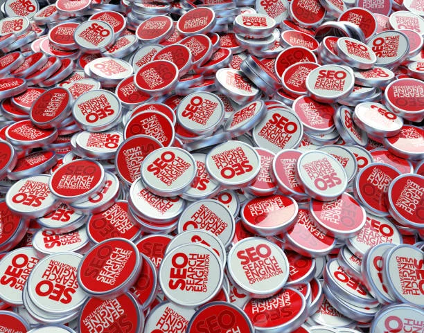 A sea of overlapping pin-back buttons, each emblazoned with the term 'SEO' in red on a white background, along with the full form 'Search Engine Optimization' in a smaller font. The buttons are densely scattered, creating a visually striking pattern of red and white. This image symbolizes the widespread importance of SEO in the digital marketing industry and the potential of tools like AdSense for monetizing content online