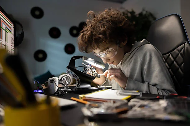 A person is intently working on an electronic project at a desk cluttered with tools and components, wearing safety goggles and using a soldering iron, illuminated by a desk lamp.