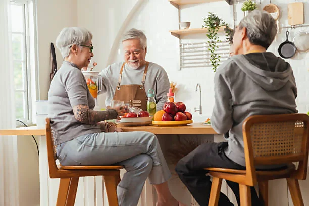 Three individuals are seated around a wooden kitchen island, engaging in conversation in a bright, cozy kitchen adorned with modern amenities and green plants. A variety of fresh fruits and beverages are placed on the island, indicating a casual gathering or meal preparation