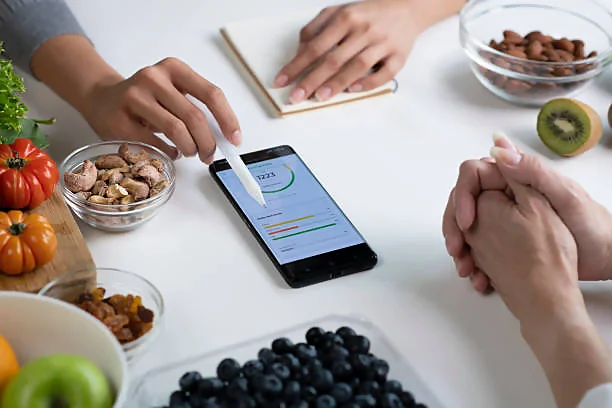 Overhead view of a person's hands interacting with a smartphone displaying a nutrition tracking app, set on a table surrounded by various healthy foods including tomatoes, almonds, raisins, and blueberries. One hand is holding a fork, while the other appears to be inputting data into the app. This image illustrates the integration of technology in monitoring daily health and nutrition