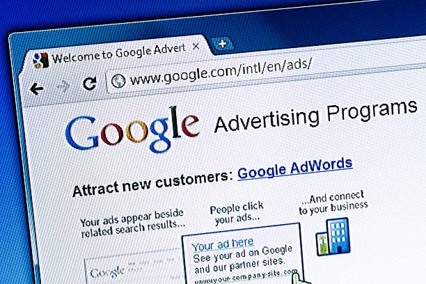 The image is a close-up of a computer monitor displaying the Google Advertising Programs webpage, with the Google logo in its traditional colors prominently at the top. The page headline reads 'Attract new customers: Google AdWords,' highlighting one of Google's advertising tools. Below this, there are two sections of text: one detailing that ads appear beside related search results, and the other emphasizing that people click the ads to connect to your business. A sample ad placement shows 'Your ad here' with a note about seeing your ad on Google and partner sites. This image communicates the essentials of using Google's advertising services to increase online revenue, relevant to the concept 'Maximize Your Online Revenue with AdSense – Start Earning Today!