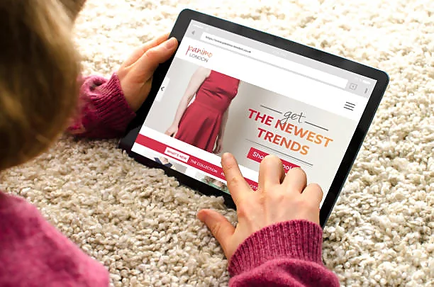 A person is seated on a plush carpet, engaging with a tablet displaying an online fashion advertisement. The ad features a large, bold title 'THE NEWEST TRENDS' alongside an image of a model wearing a stylish red skirt. The user's fingers are poised to tap a 'Shop Now' button, suggesting interaction with the banner ad. This setting conveys how banner advertisements on websites can entice visitors to explore products and potentially increase website revenue through user engagement