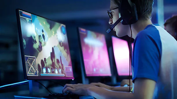 A person in a blue shirt and wearing headphones is intensely focused on playing an online game, depicted on a brightly lit computer monitor, in a dark room illuminated by the screens’ glow.