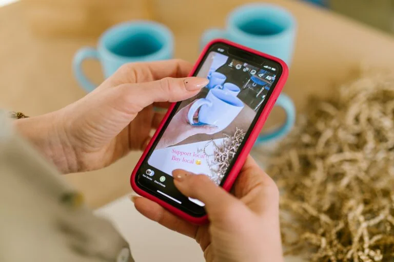 A person’s hands holding a smartphone with a red case, capturing an image of three blue mugs on a table. The phone screen displays the photo being taken, with text encouraging support for local businesses. The setting includes some decorative dried plants, emphasizing the theme of local community support in the context of large-scale advertising campaigns.