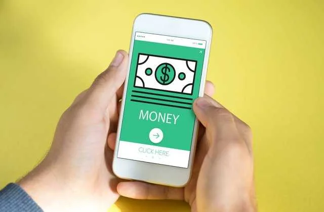 A person holding a smartphone displaying an app screen with a simple graphic of a dollar bill and the word 'MONEY' in bold green letters. Below the text is a button labeled 'CLICK HERE'. The phone is positioned against a plain yellow background, suggesting an interface for an online money-making or financial app